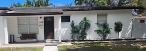 A single-story house with a white exterior, a dark brown door, and a number '1805' on the front wall. There is a small porch with a bench and a trash can. - Pool Home w/ 2 Kings near Beach, FLL & Hard Rock (Hollywood)