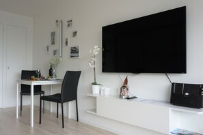 A modern living room with a flat-screen TV mounted on the wall, a dining table with chairs, and a small white shelf with decorative items. - New Listing (Davos Platz)