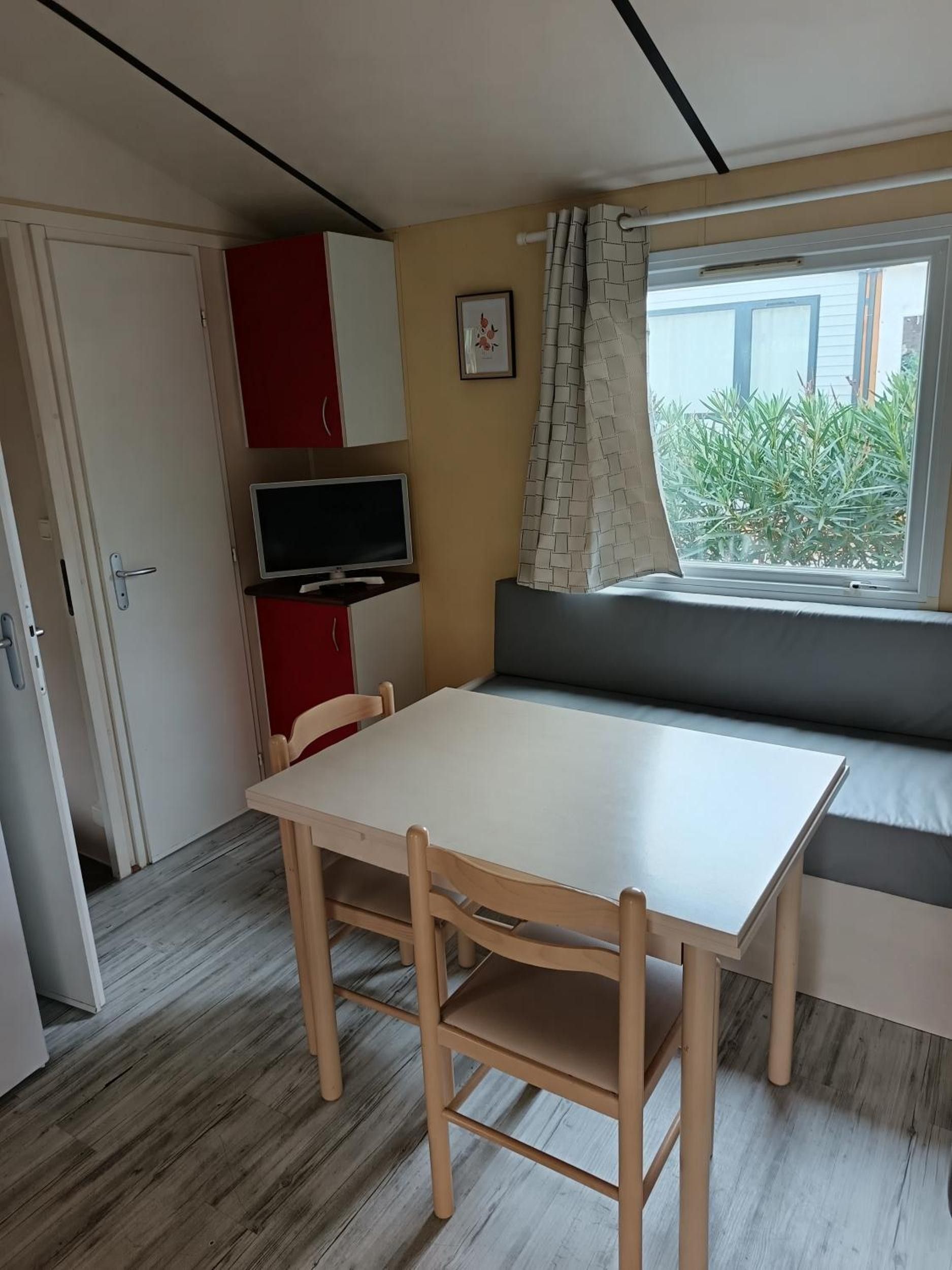 Une pièce avec une table et des chaises blanches, un meuble rouge, un téléviseur, une fenêtre donnant sur un paysage verdoyant et une porte.