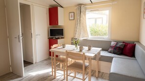 A compact room with a dining table, chairs, a sofa, a TV, and a window with curtains. - Mobile Home 'Camping Le Clos Du Thym' with Shared Pool, Wi-Fi and Air Conditioning (Argelès-sur-Mer)