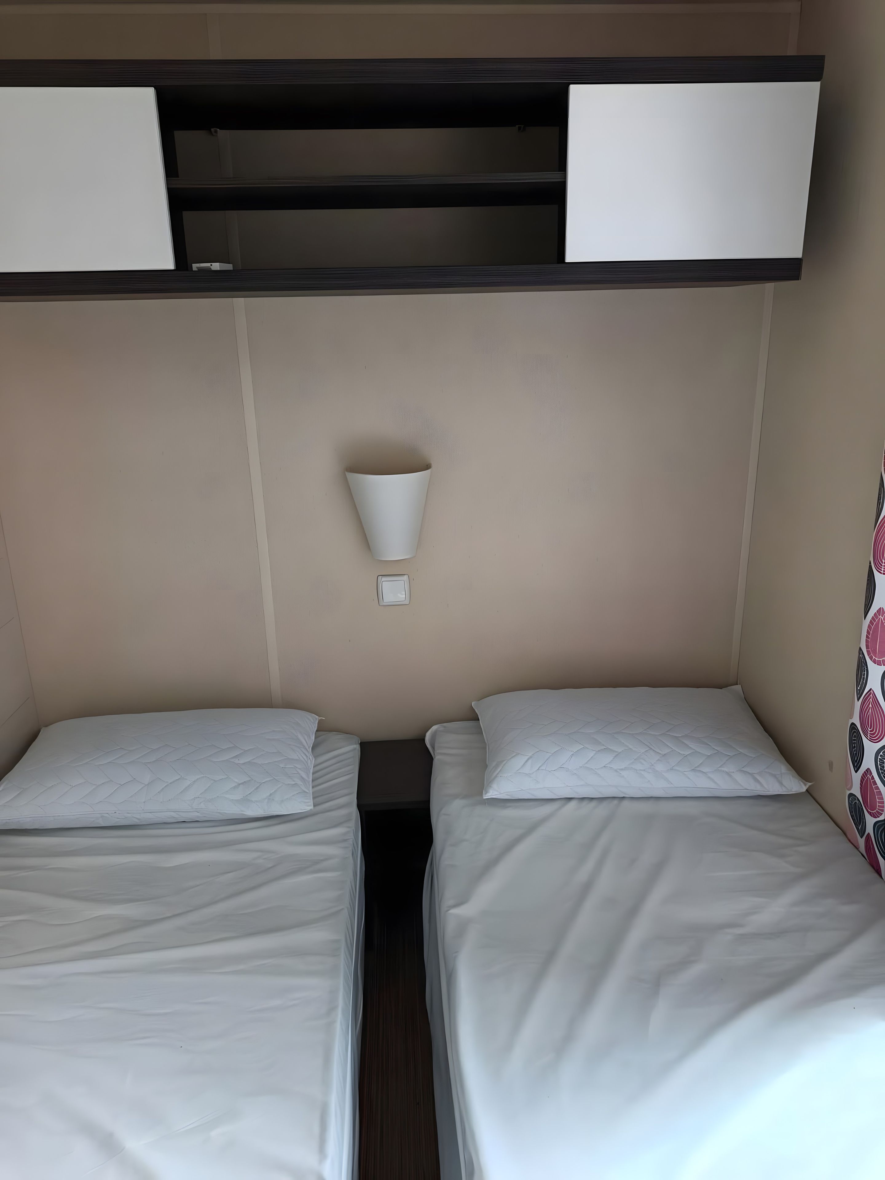 Een kamer met twee bedden, een klein schapje erboven en een wandlamp.