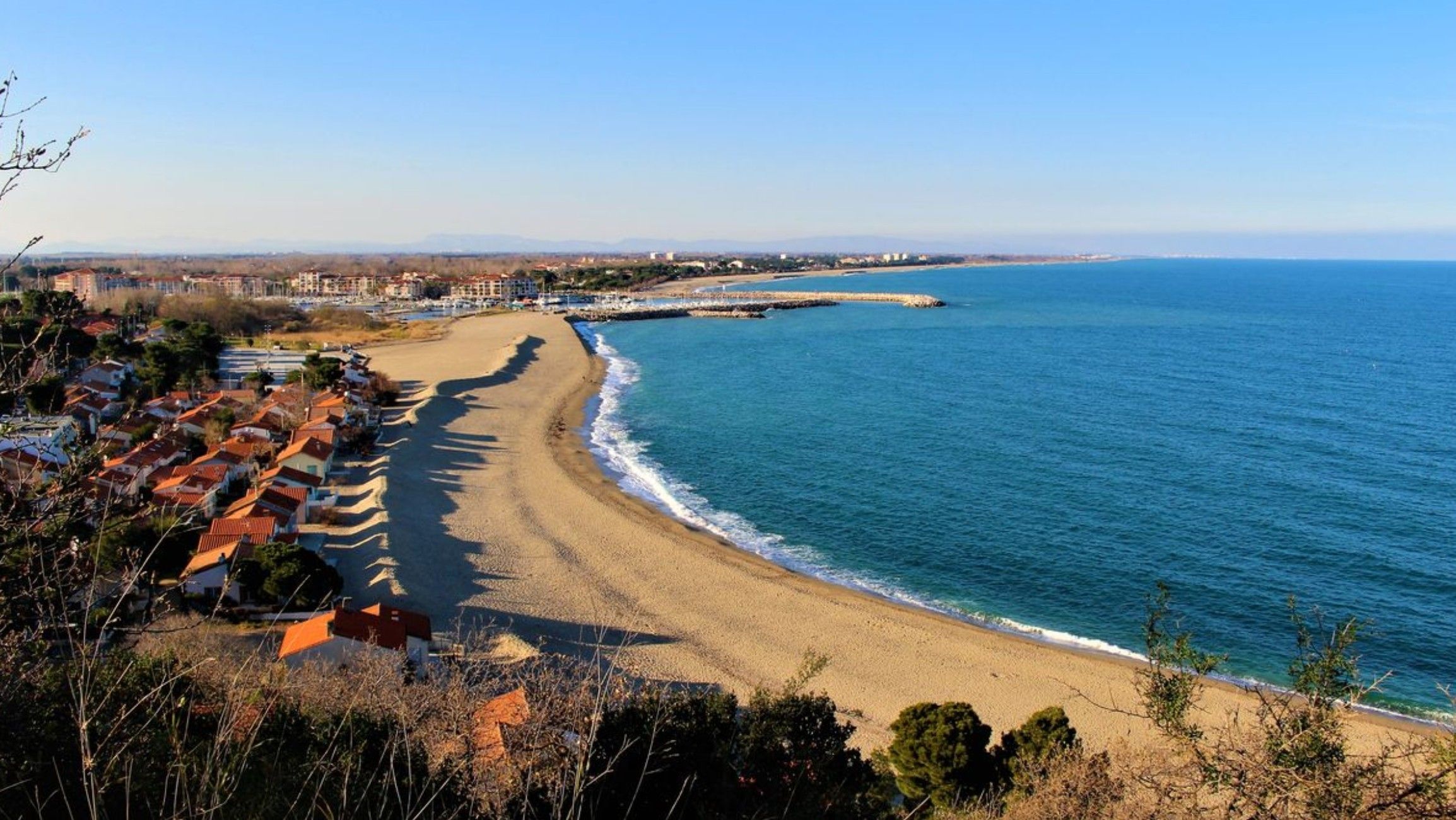 A coastal view with a sandy beach, a clear blue sea, and a row of buildings with red roofs.