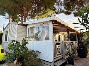 A mobile home with a covered porch, a car parked in front, and various plants around. - Mobile Home 'Avec Piscine' with Shared Pool, Wi-Fi and Air Conditioning (Argelès-sur-Mer)