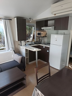 A compact kitchenette with a microwave, stove, and refrigerator. There is a dining table with chairs and a sofa in the living area. - Mobile Home 'Avec Piscine' with Shared Pool, Wi-Fi and Air Conditioning (Argelès-sur-Mer)