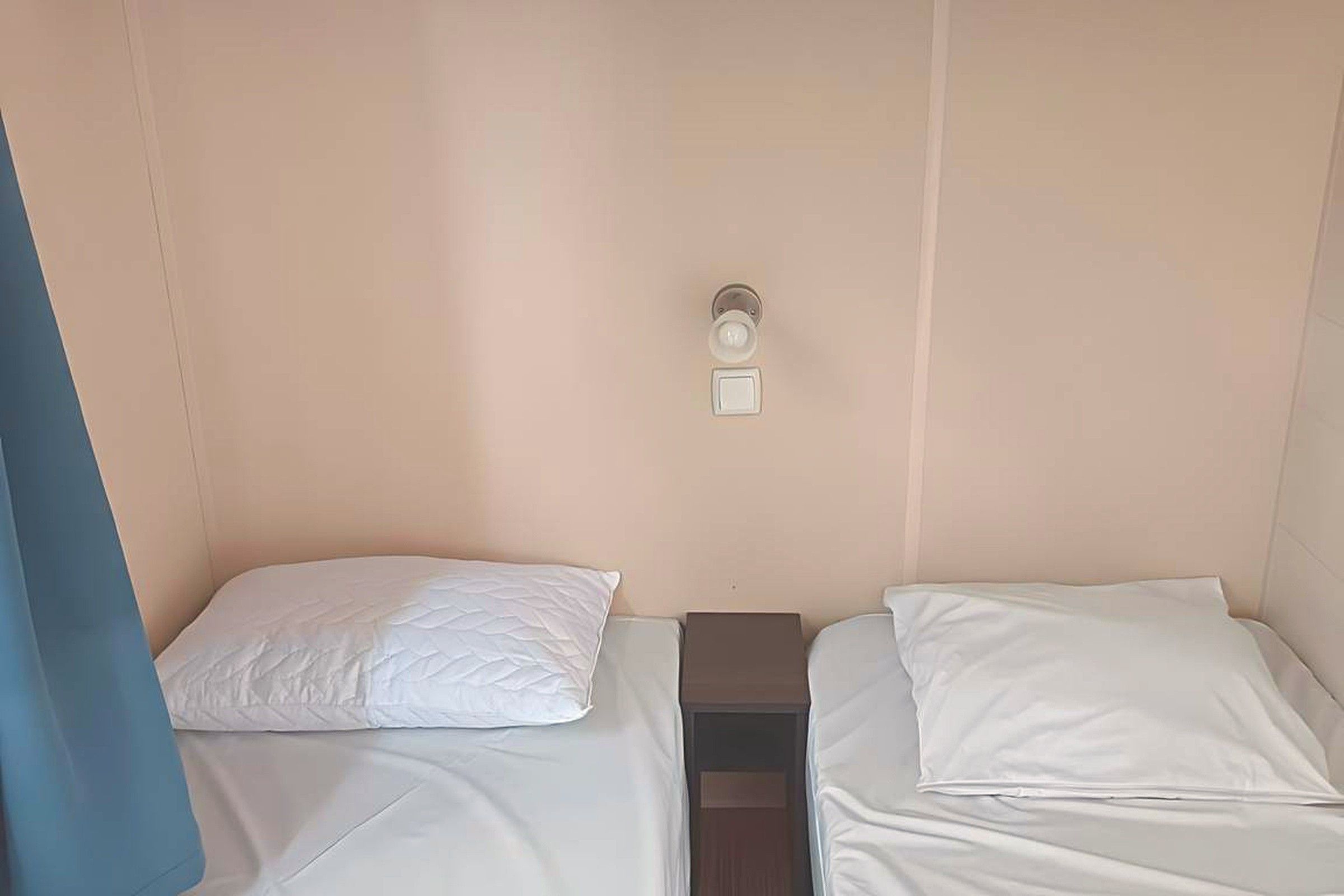 Un petit logement composé d’une seule pièce, comprenant deux lits, une table de chevet et un luminaire fixé au mur.