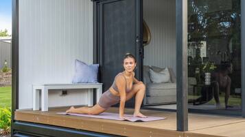 Une personne effectue une posture de yoga sur un tapis, à l’extérieur d’un bâtiment moderne doté d’une porte en verre, d’un banc et d’un canapé à l’intérieur.
