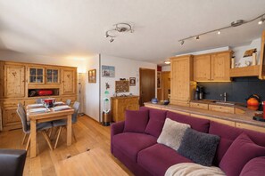 A cozy living area with a wooden dining table, a purple sofa, and a kitchen with wooden cabinets and a coffee machine. - App. La Forêt 43 - Two Bedroom Apartment, Sleeps 5 (Riddes)