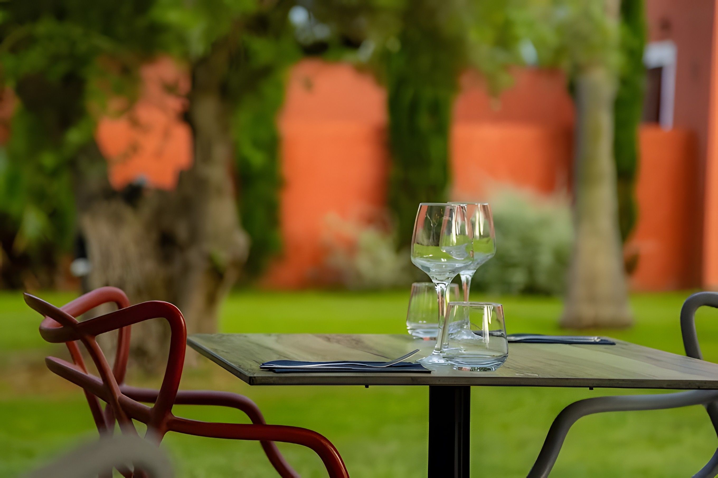 Une table avec deux verres à vin et un verre à eau, placée à l’extérieur, avec une pelouse verte et un mur rouge en arrière-plan.