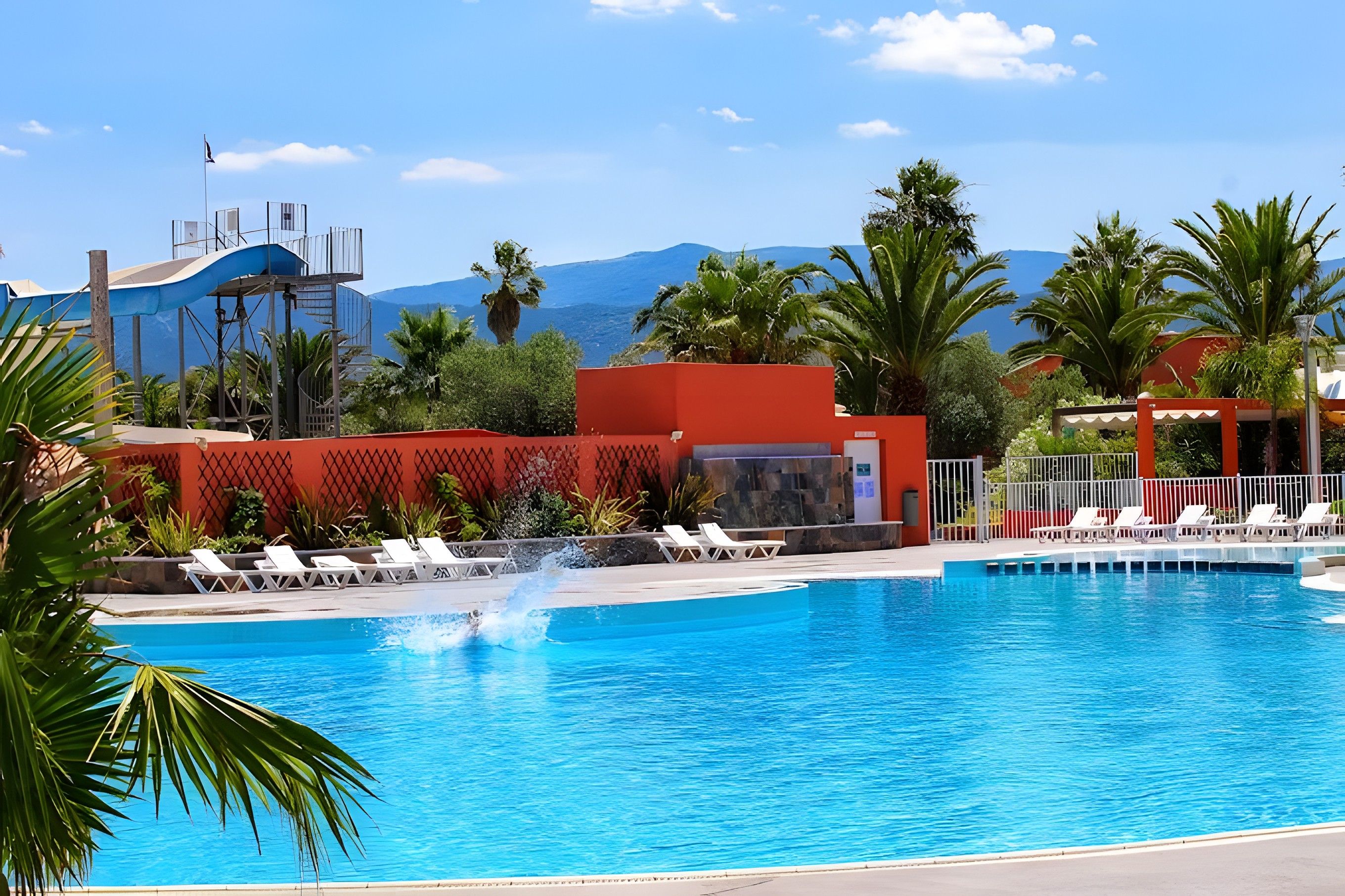 A swimming pool with clear blue water, surrounded by palm trees and a red building.
