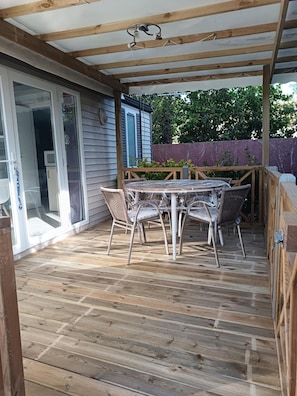 A covered patio with a wooden table and wicker chairs, a wooden deck, and a sliding glass door leading to a room. - Mobile Home 'Mobil-home Piscine, Clim Et Wifi' with Shared Pool, Wi-Fi and Air Conditioning (Argelès-sur-Mer)