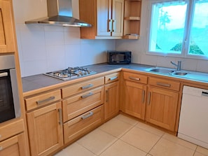 A kitchen with wooden cabinets, a gas stove, a microwave, a sink, and a window with a view of palm trees. - Sea-view Holiday Home in Hanvec for 5 (Hanvec)