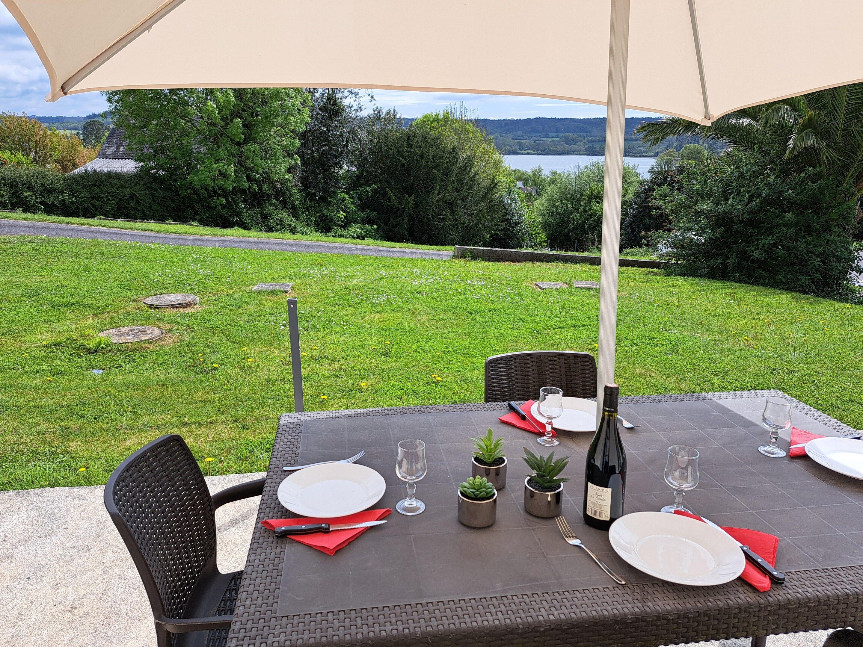 An outdoor dining table set with plates, cutlery, and wine glasses, surrounded by chairs, with a view of a grassy area and trees in the background.