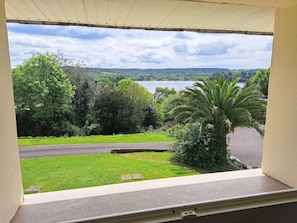 A view from a window overlooking a grassy area with a palm tree, a road, and a lake in the distance. - Sea-view Holiday Home in Hanvec for 5 (Hanvec)