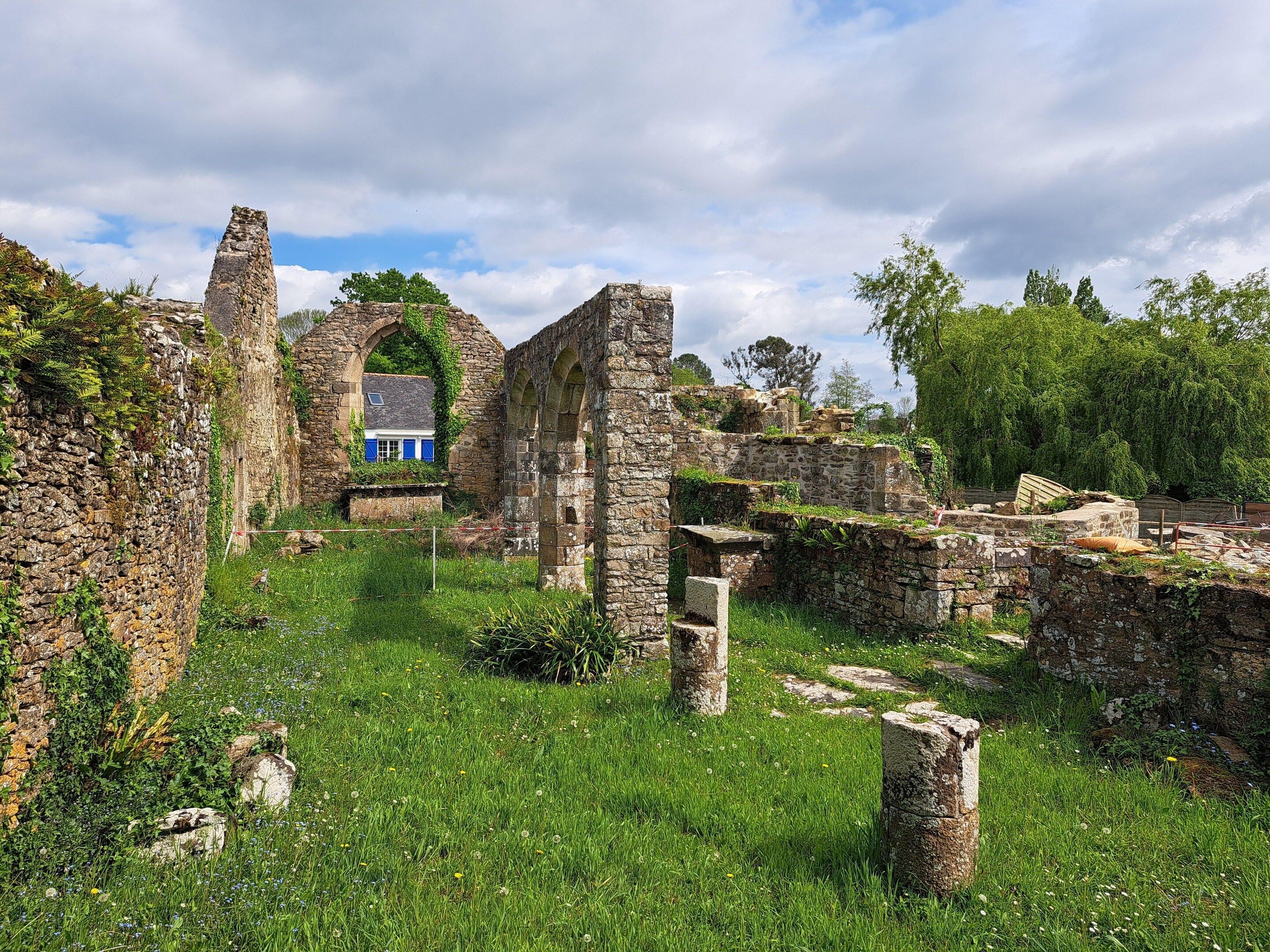 Ancient stone ruins with overgrown vegetation, a small building with blue shutters, and a clear sky.