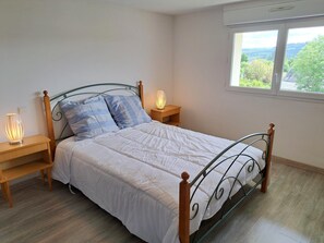 A neatly made bed with a metal frame and wooden headboard, flanked by bedside tables with lamps, in a room with a large window offering a view of greenery. - Sea-view Holiday Home in Hanvec for 5 (Hanvec)
