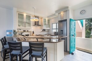 A modern kitchen with a central island, built-in cabinets, and a dining area with chairs. - Santorini Ballito | 180º Sea Views, Braai and Pool (Cape Town)