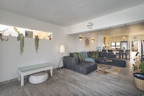A modern living room with a grey sofa, a glass coffee table, and a white bench. There is a kitchen visible in the background. - Santorini Ballito | 180º Sea Views, Braai and Pool (Cape Town)
