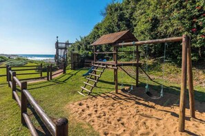 A playground with a wooden swing set, a slide, and a climbing structure. - Santorini Ballito | 180º Sea Views, Braai and Pool (Cape Town)