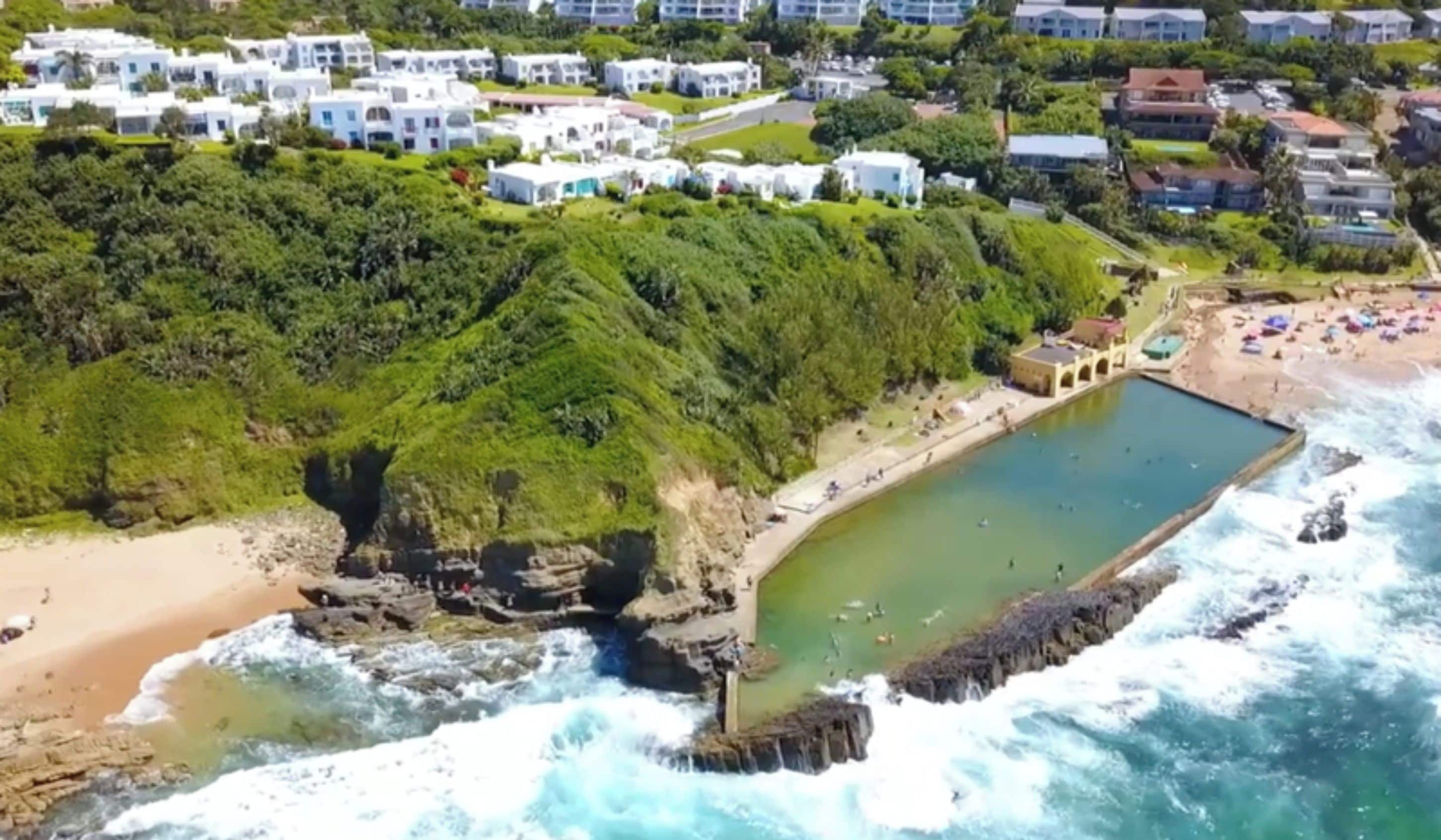 A coastal area with a rocky beach, a swimming pool, and a residential area with white buildings.