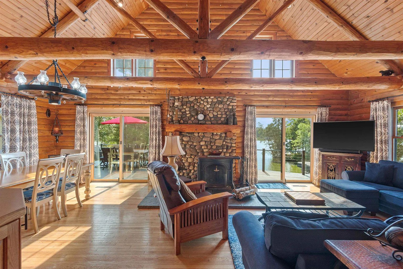 A log cabin interior with a fireplace, wooden furniture, and a large window overlooking a body of water.