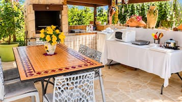 An outdoor patio with a stone floor, a table with a patterned tablecloth, a chair, a potted plant with yellow flowers, and a barbecue grill.