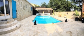 A swimming pool with stone steps leading to it, a wooden building with a blue door, and a small statue on the pool's edge.
