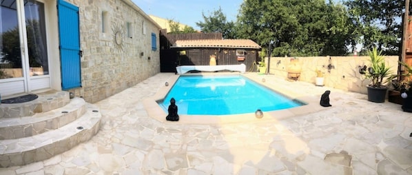 Une piscine avec des marches en pierre menant à l’eau, un bâtiment en bois avec une porte bleue et une petite statue placée au bord de la piscine.