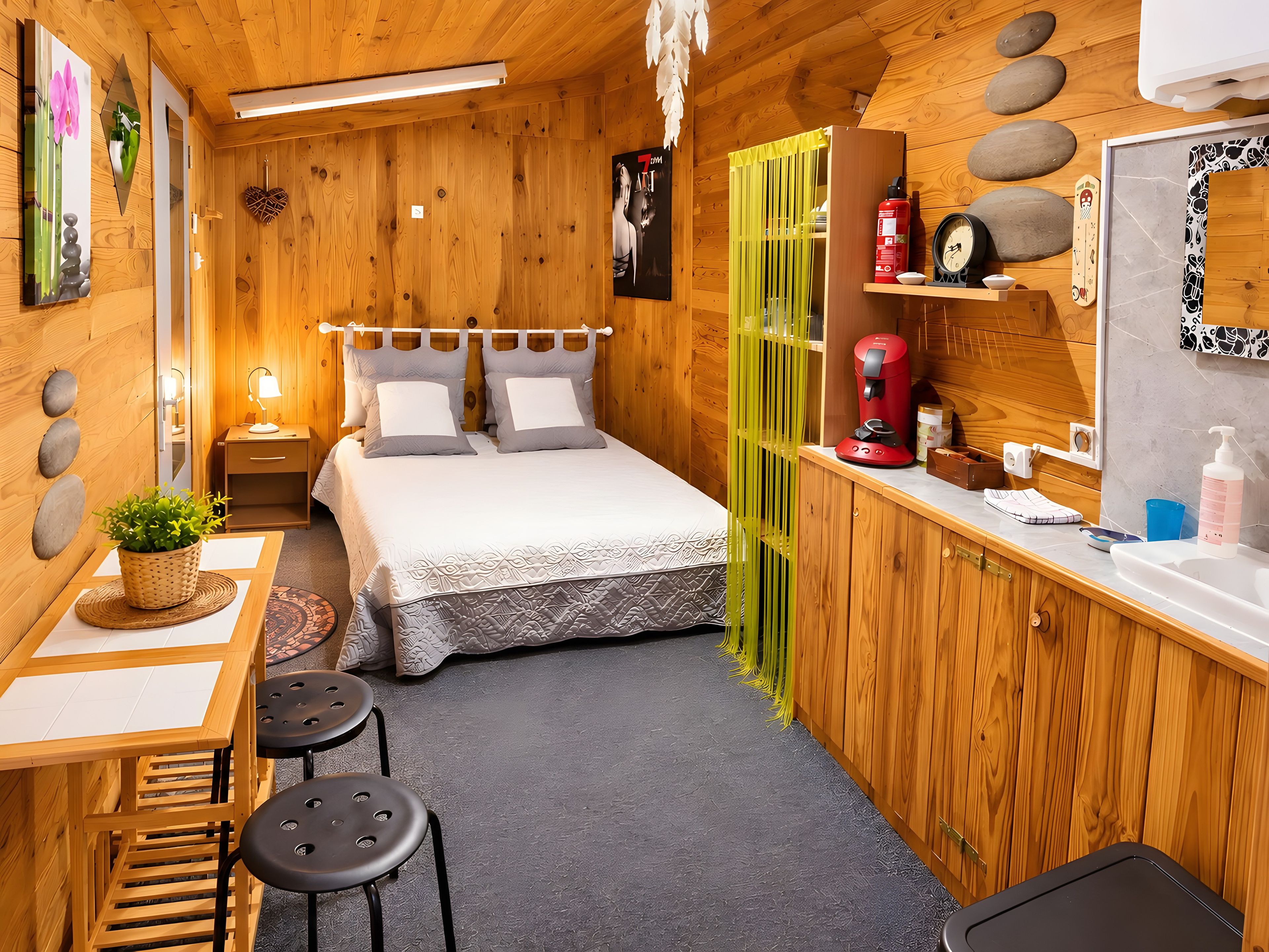 A wooden interior with a bed, bedside table, lamp, and a small desk with a chair.
