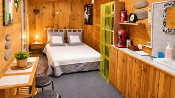 A wooden interior with a bed, bedside table, lamp, and a small desk with a chair.