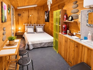 A wooden interior with a bed, bedside table, lamp, and a small desk with a chair.