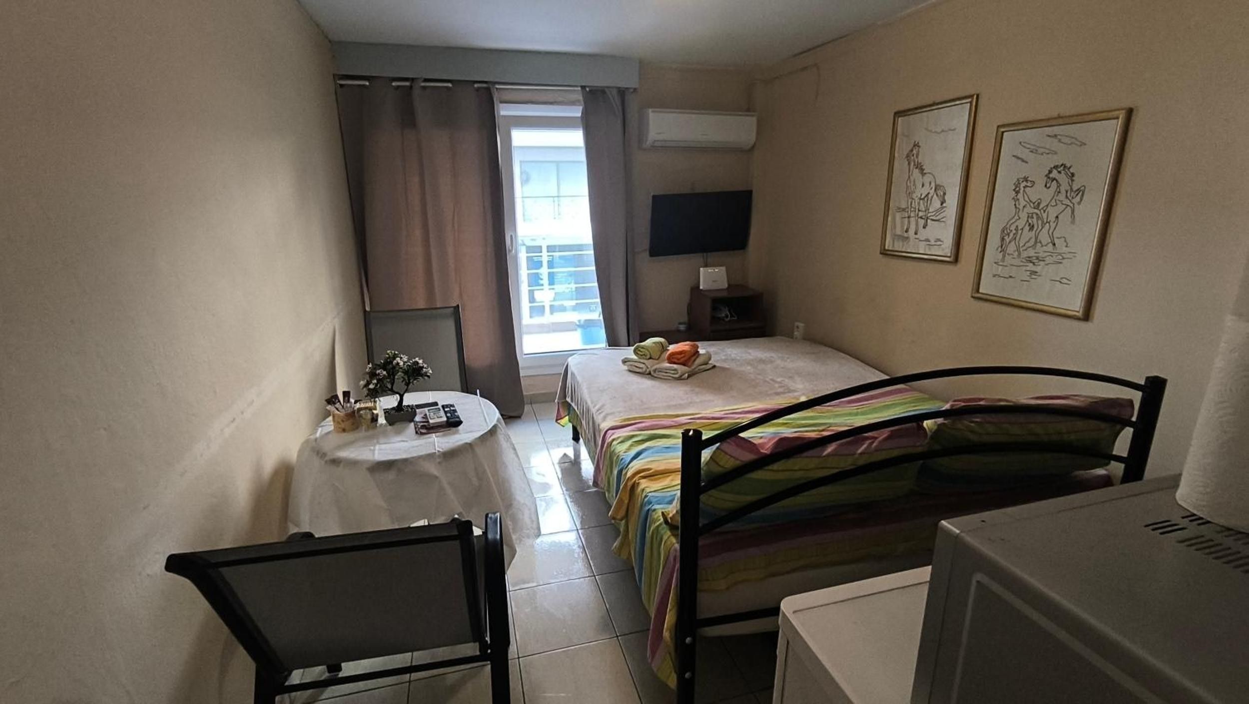 Ein kleines Hotelzimmer mit einem Bett, einem Schreibtisch, einem Sessel, einem Fernseher und Bildern an den Wänden.
