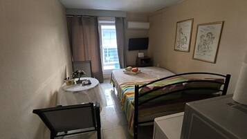 Ein kleines Hotelzimmer mit einem Bett, einem Schreibtisch, einem Sessel, einem Fernseher und Bildern an den Wänden.