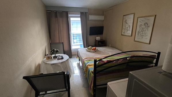 Ein kleines Hotelzimmer mit einem Bett, einem Schreibtisch, einem Sessel, einem Fernseher und Bildern an den Wänden.