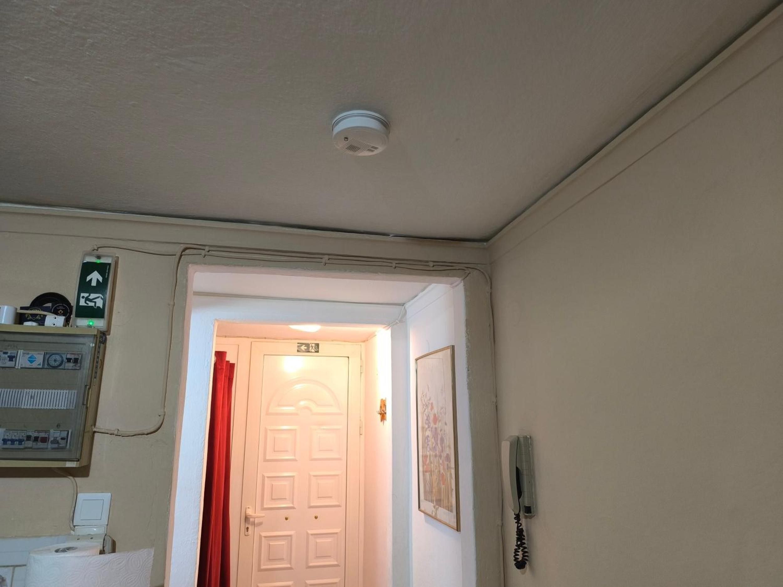 Une pièce avec une porte blanche, un rideau rouge, un téléphone fixé au mur et un détecteur de fumée fixé au plafond.