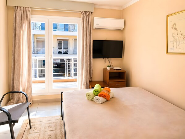 A hotel room with a bed, a TV, a window with curtains, a small wooden nightstand, and a framed picture on the wall.
