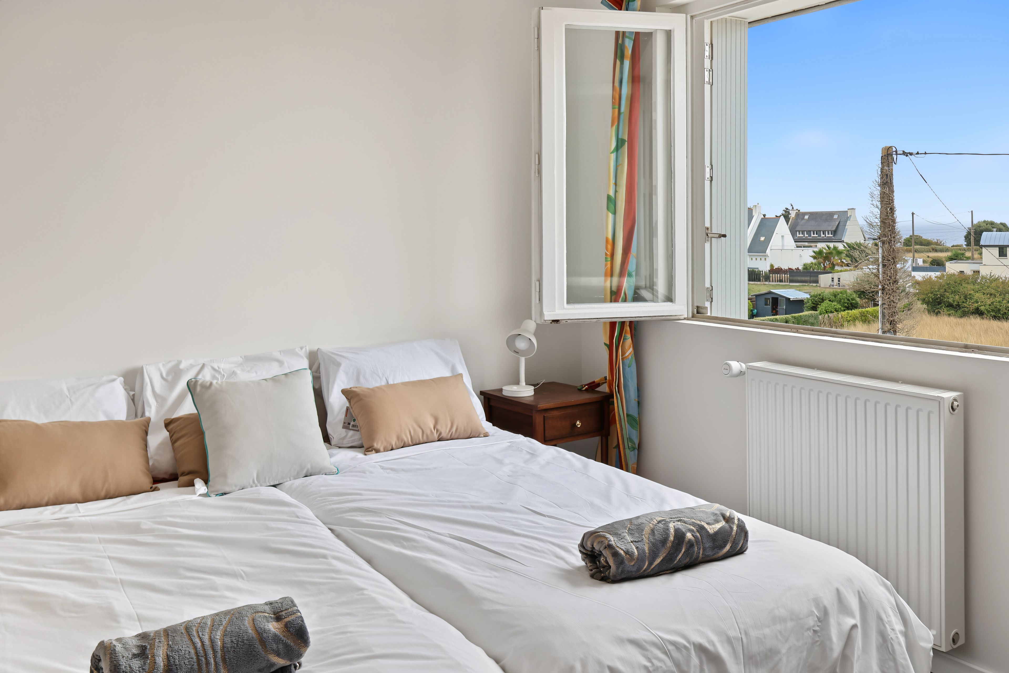 A bedroom with a bed, pillows, a bedside table, a lamp, a window with curtains, and a view of the outside.