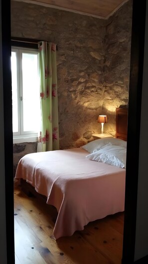 A bedroom with a stone wall, a bed with a pink bedspread, a nightstand with a lamp, and a window with curtains.