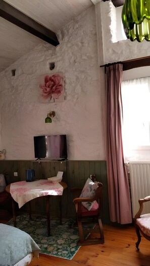 A room with a wooden floor, a television on a stand, a table with a floral tablecloth, a chair with a floral cushion, and a large pink flower decoration on the wall.