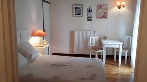 Ein Schlafzimmer mit einem Bett, einem Nachttisch mit Lampe, einer Heizung, einem weißen Stuhl, einem kleinen Tisch und Wandbildern.