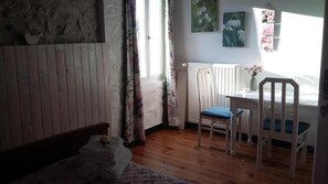 A room with a wooden floor, a bed with a quilt, a chair with a cushion, a radiator, and a window with curtains.