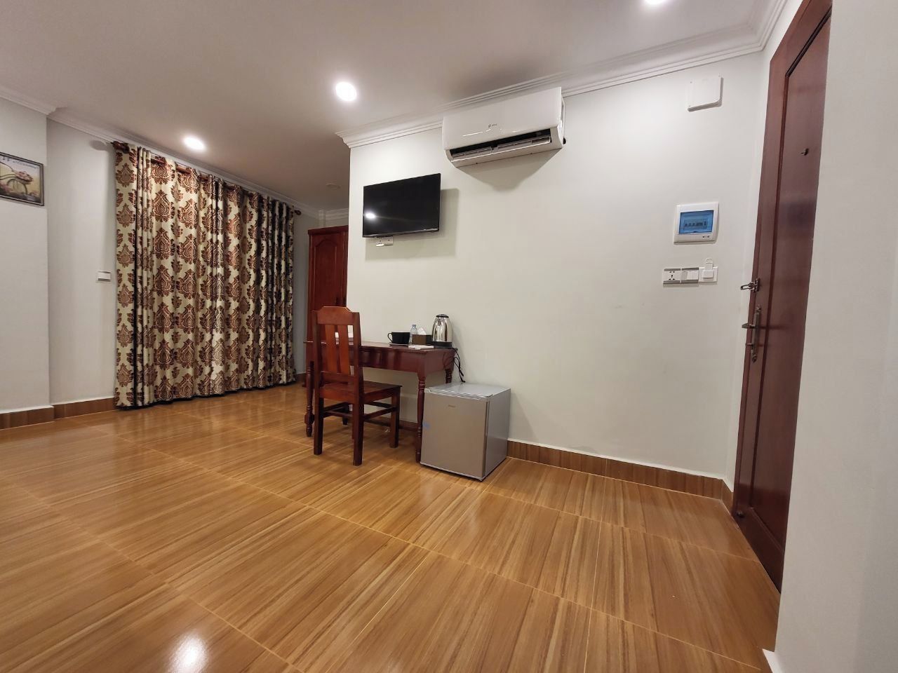 A room with a wooden floor, a TV mounted on the wall, a small refrigerator, and a door.