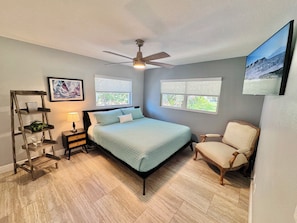 Quarto com cama, cadeira, mesa de cabeceira, luminária, ventilador de teto e duas janelas.