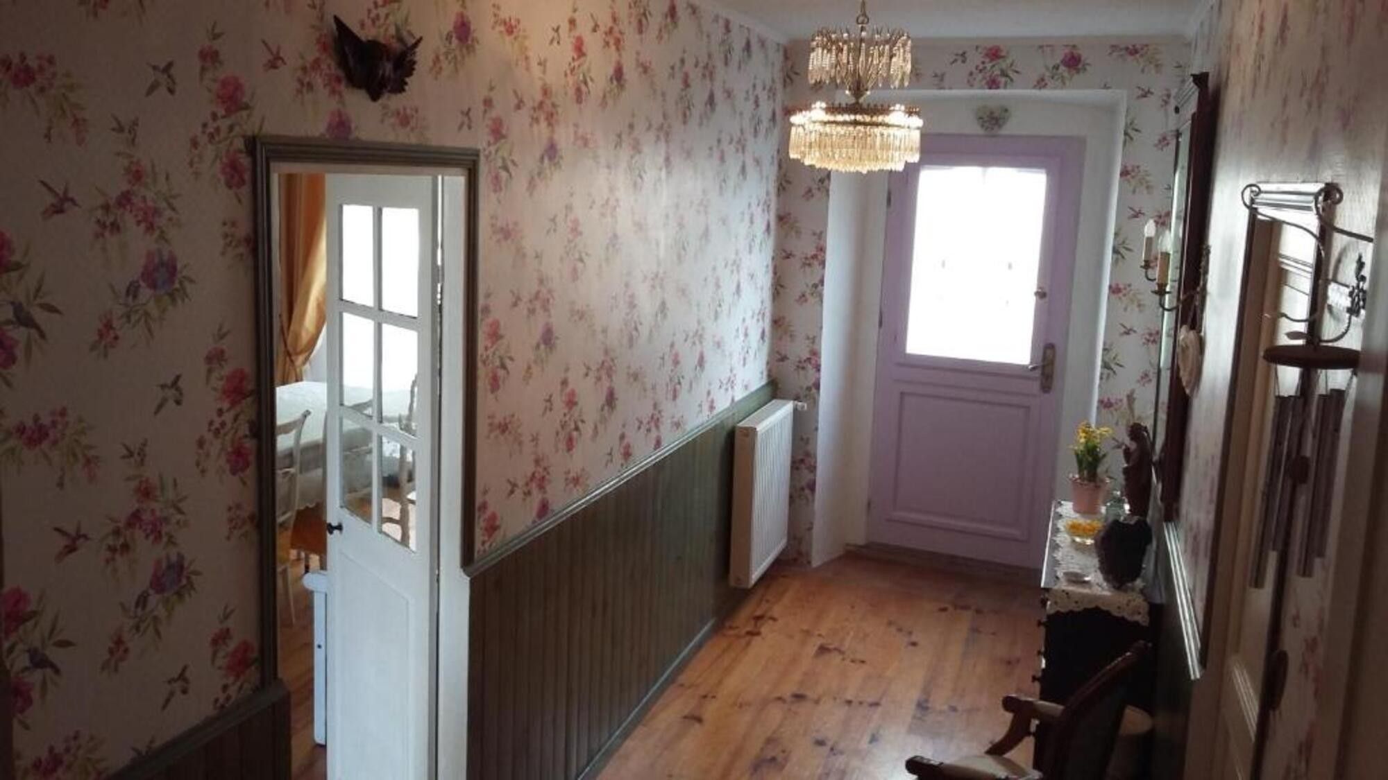 Un couloir avec un papier peint à motifs floraux, un plancher en bois, un lustre et une porte dotée d’une fenêtre.