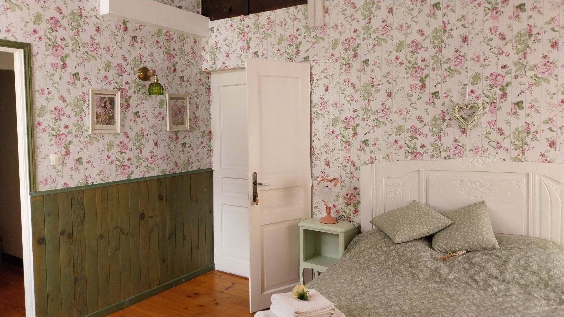 Un dormitorio con papel tapiz floral, suelo de madera, cama con ropa de cama verde, mesita de noche y una puerta.