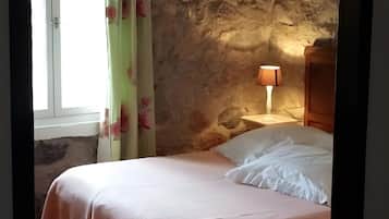 A bedroom with a stone wall, a bed with a pink bedspread, a nightstand with a lamp, and a window with curtains.