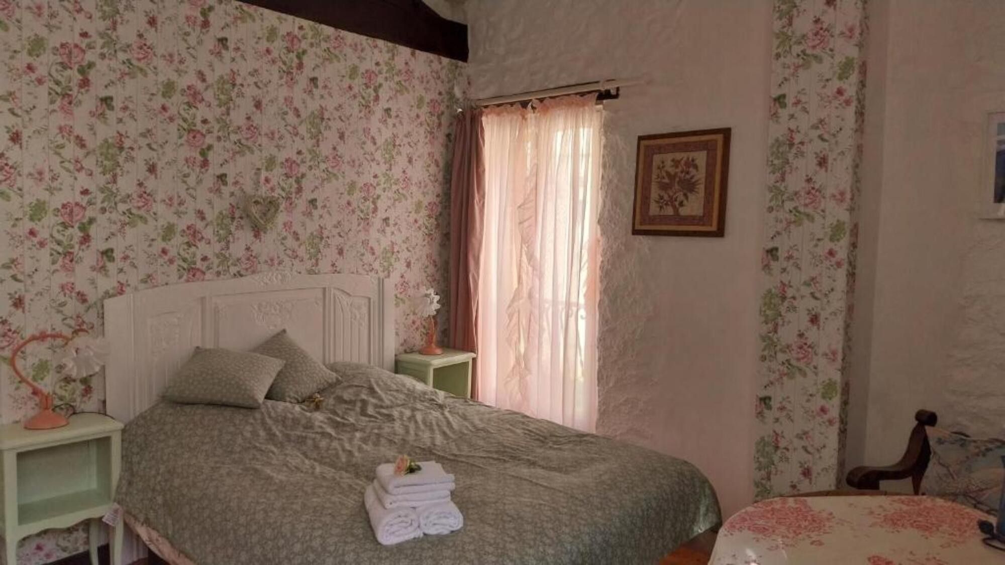 Une chambre à coucher avec un papier peint à motifs floraux, un lit avec des oreillers, une table de chevet, une fenêtre avec des rideaux et un tableau encadré accroché au mur.
