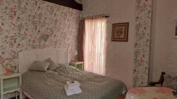 Une chambre à coucher avec un papier peint à motifs floraux, un lit avec des oreillers, une table de chevet, une fenêtre avec des rideaux et un tableau encadré accroché au mur.