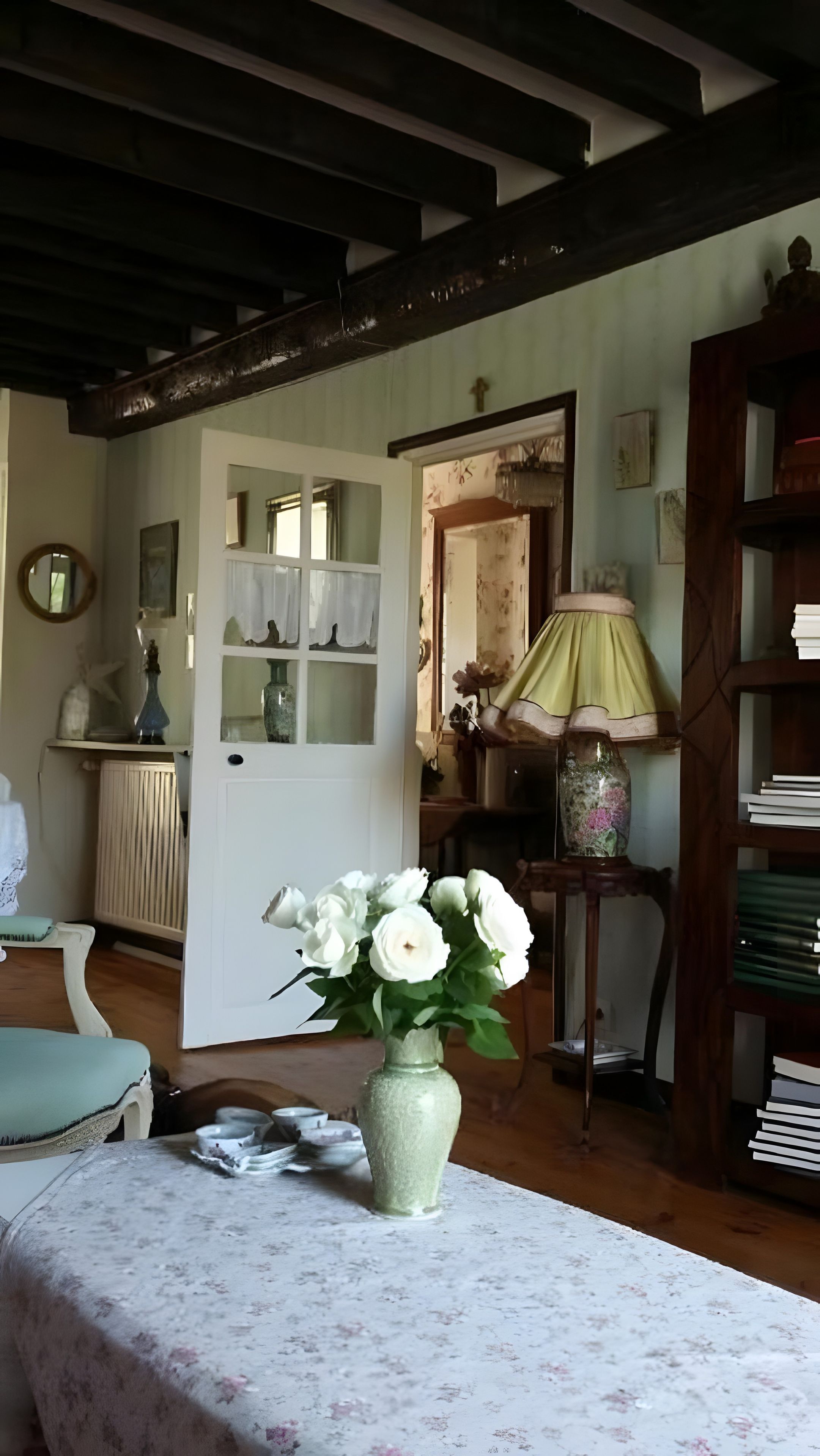 A room with a table, a vase with flowers, a lamp, and a bookshelf.