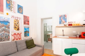 A modern living room with a grey sofa, a green pillow, and a kitchen area with a sink, a red microwave, and various kitchen items. - New Listing (Napoli)