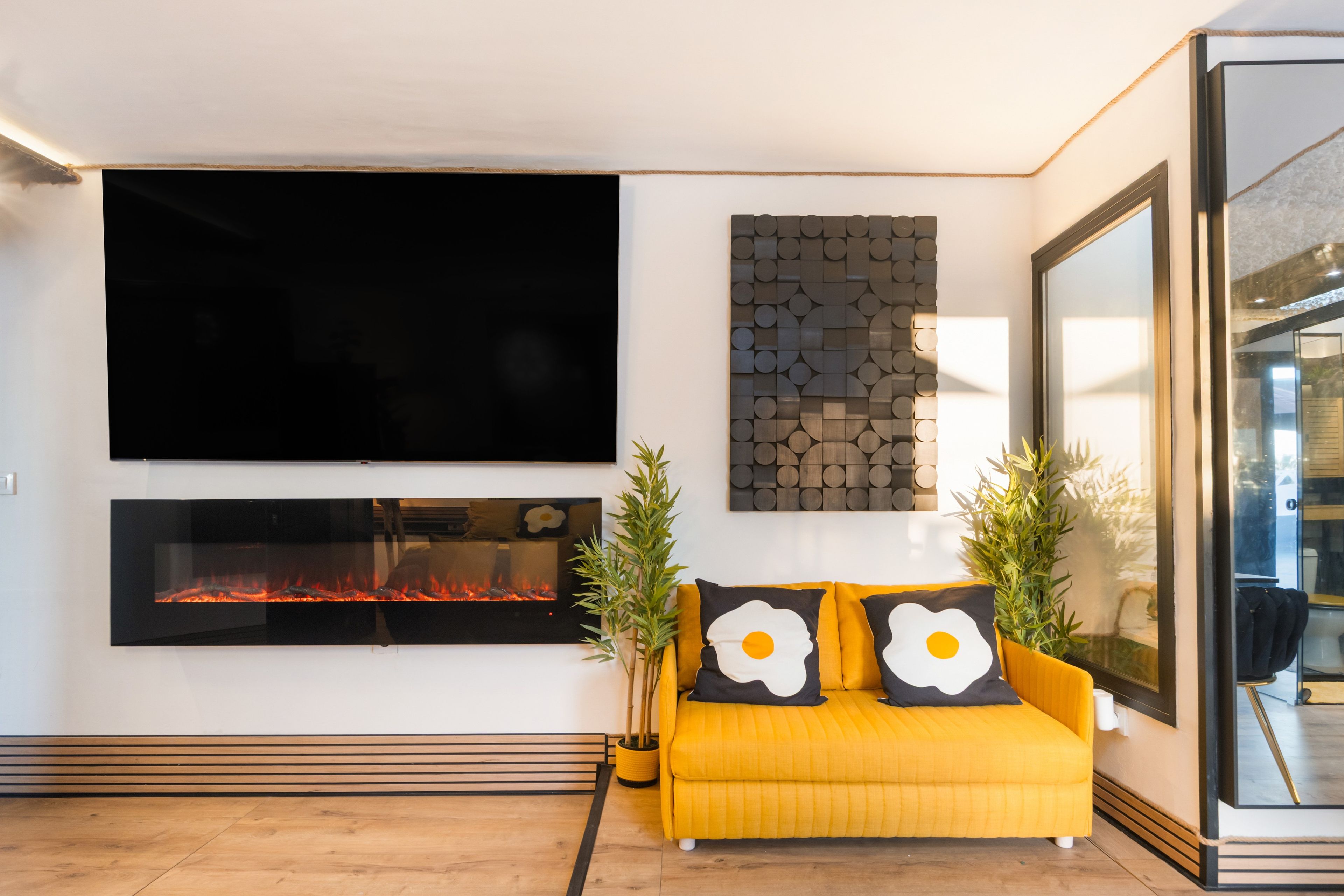 A modern living room with a flat-screen TV, a fireplace, a yellow sofa with decorative pillows, and a wall-mounted artwork.